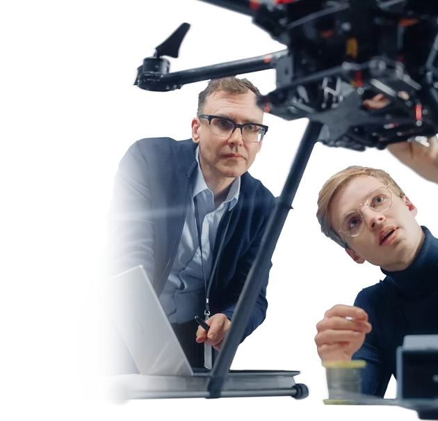 Zwei M&auml;nner pr&uuml;fen und besprechen technische Details an einer Drohne, w&auml;hrend ein Laptop auf dem Tisch steht.