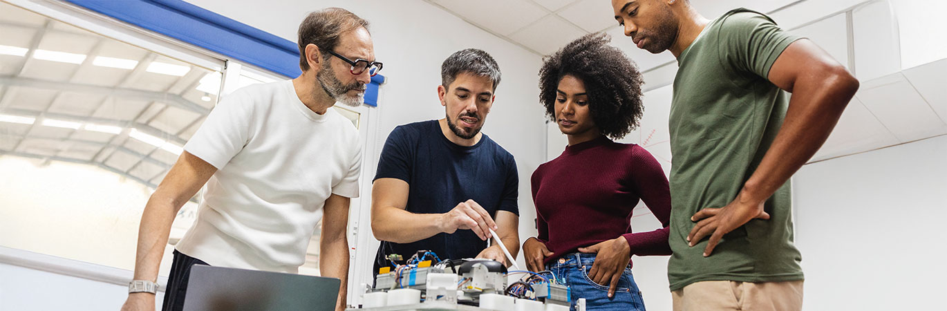 Eine Gruppe von Ingenieuren und Ingenieurinnen arbeitet gemeinsam an einem elektronischen Prototypen auf einem Tisch im Labor.