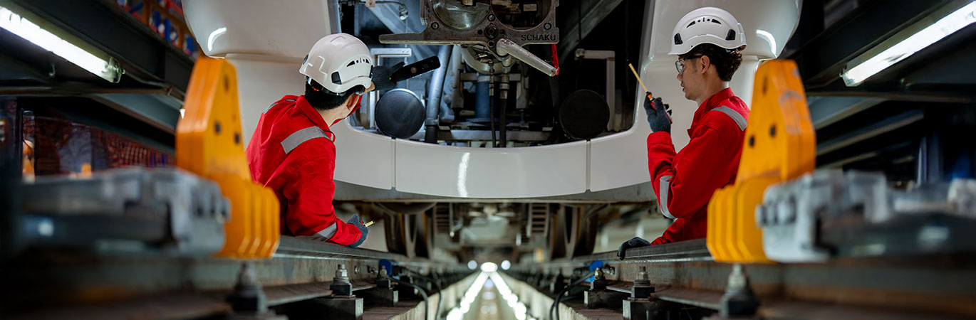 Zwei Ingenieure in roter Schutzkleidung und wei&szlig;en Helmen &uuml;berpr&uuml;fen den Unterboden eines Zuges in einer Wartungshalle.