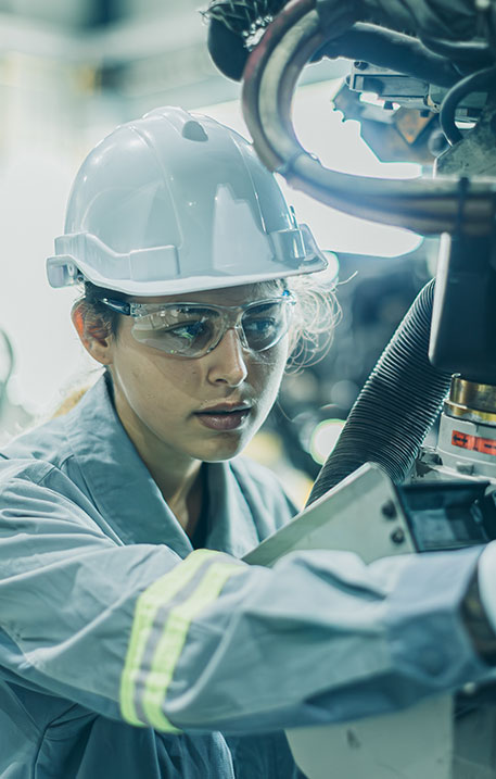 Eine Ingenieurin mit Schutzhelm und Schutzbrille arbeitet konzentriert an einer Maschine in einer Werkstatt.