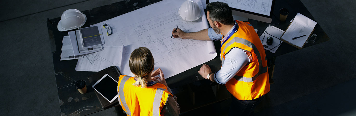 Zwei Projektingenieure mit Warnwesten und Baupl&auml;nen arbeiten gemeinsam an einem Tisch und besprechen Details einer technischen Zeichnung.