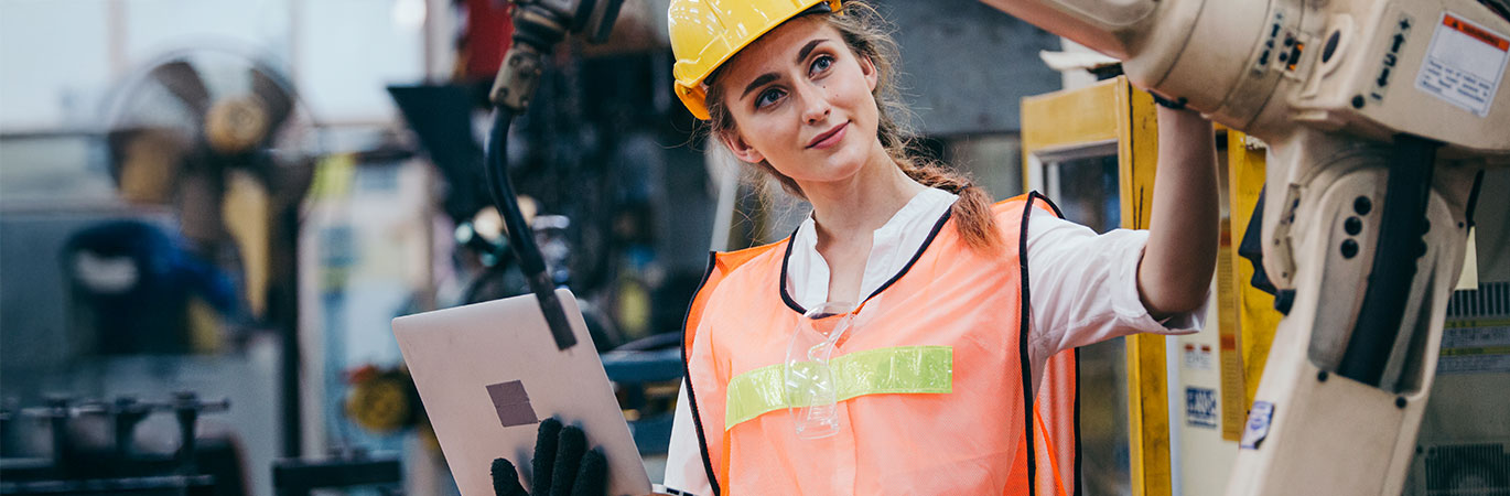 Eine Ingenieurin mit Schutzhelm, Warnweste und Laptop inspiziert eine Fertigungsanlage und &uuml;berpr&uuml;ft einen Industrieroboter.