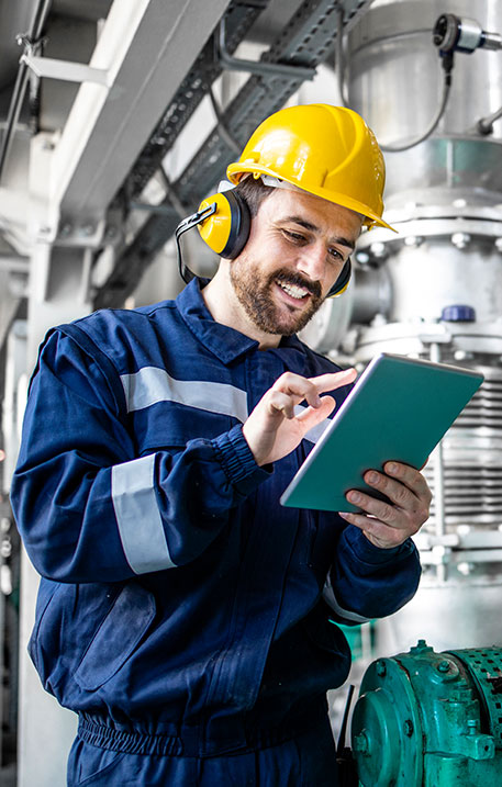 Ein Ingenieur mit gelbem Schutzhelm und Geh&ouml;rschutz bedient ein Tablet zur Qualit&auml;tskontrolle in einer Industrieanlage.