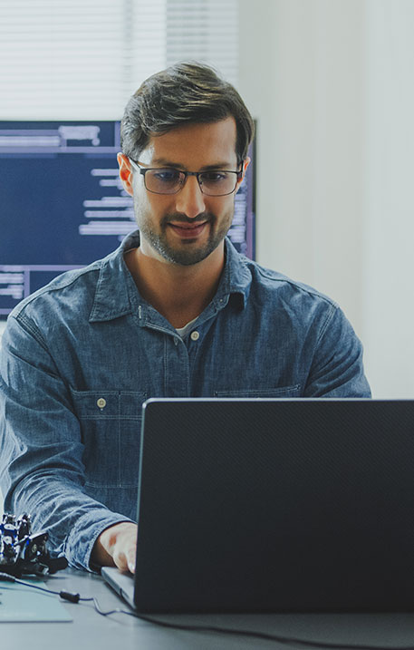 Ein Ingenieur mit Brille sitzt vor einem Laptop und entwickelt Software, im Hintergrund ist Programmcode auf einem Bildschirm sichtbar.