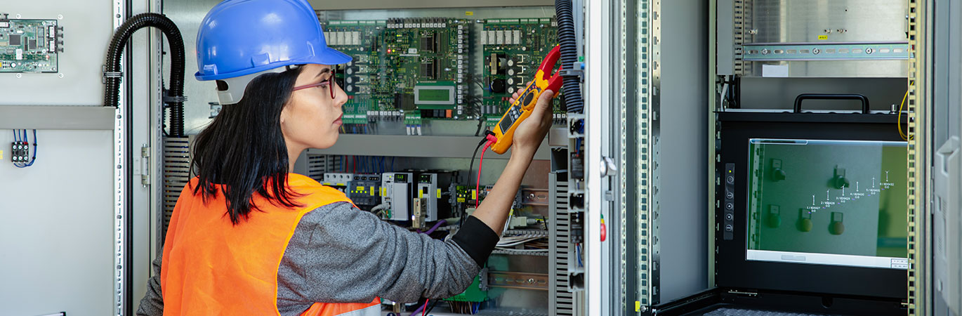 Eine Ingenieurin mit Schutzhelm und Warnweste misst mit einem Pr&uuml;fger&auml;t die Elektronik in einem Schaltschrank.